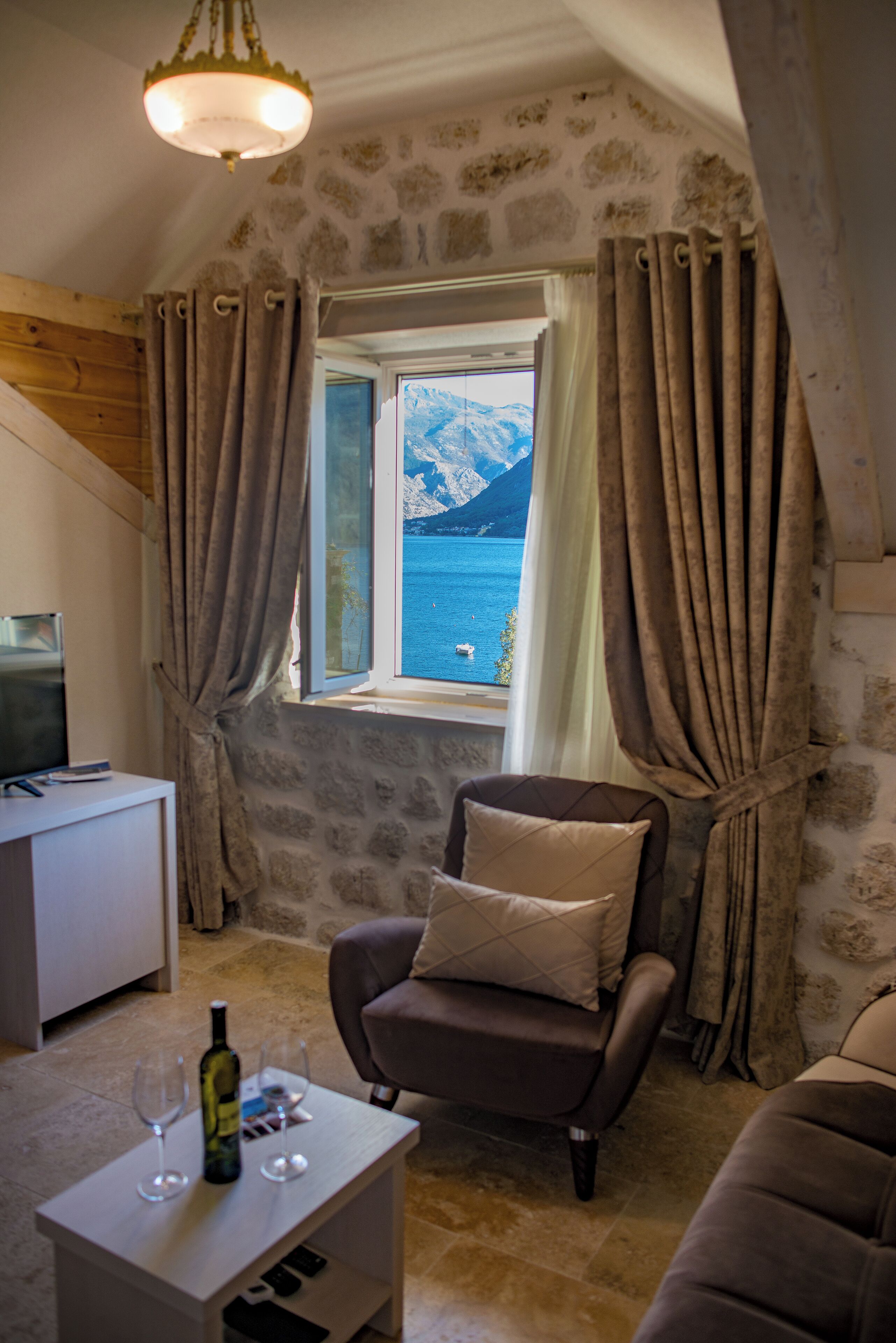 A cozy room with a stone wall, a view of a lake and mountains, a brown leather armchair, a small white table with a bottle and glasses, and a television on a white cabinet.