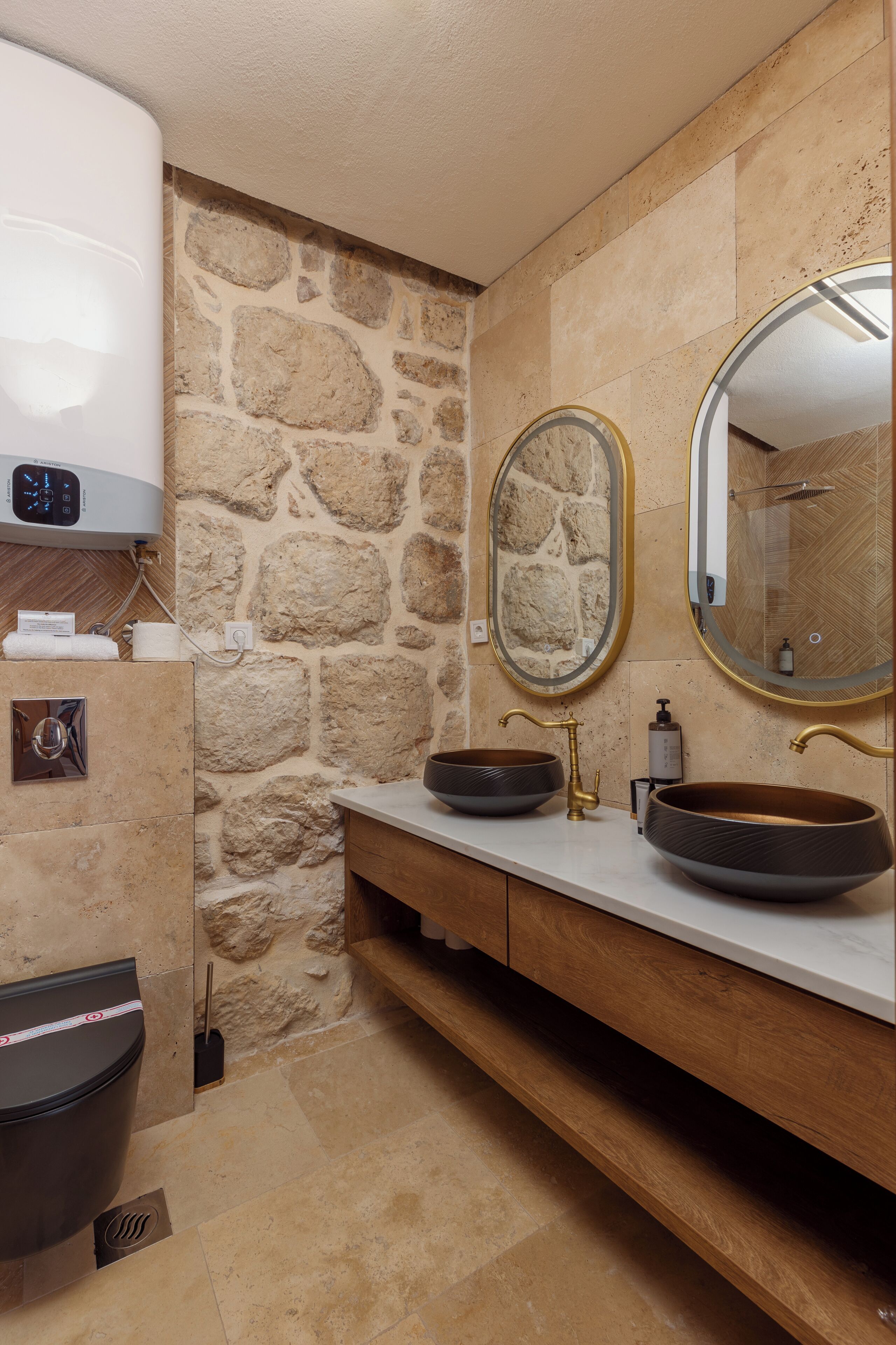 A bathroom with a stone wall, a built-in heater, a toilet, a sink with a large mirror, and a shower area.