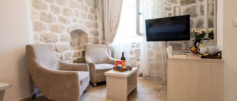 A cozy living room with stone walls, two beige armchairs, a small wooden coffee table with a bottle and snacks, a flat-screen TV, and a wooden side table with a vase and books.
