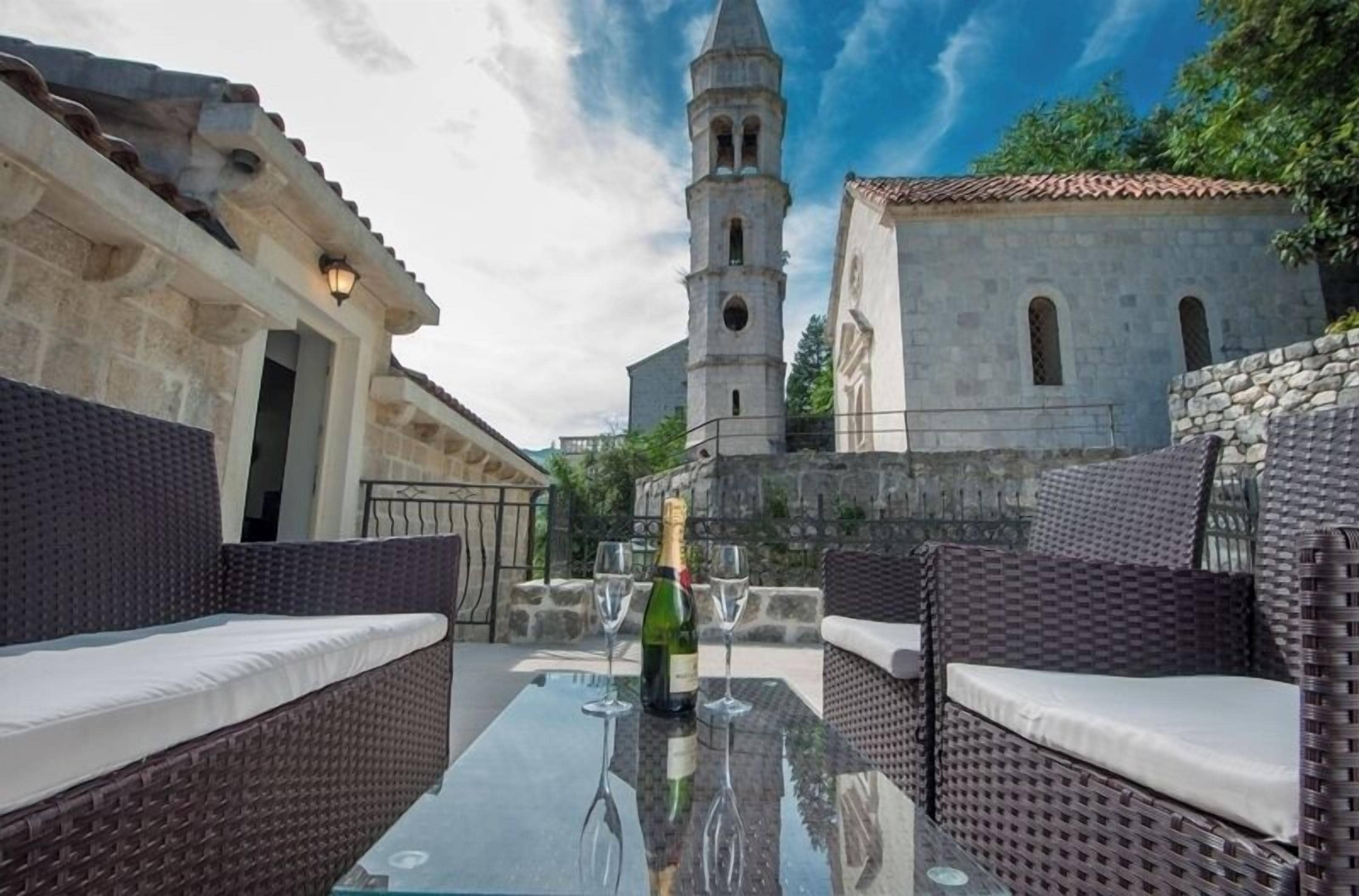 An outdoor seating area with wicker furniture, a glass table, and a bottle of champagne. A historic building with a bell tower is visible in the background.