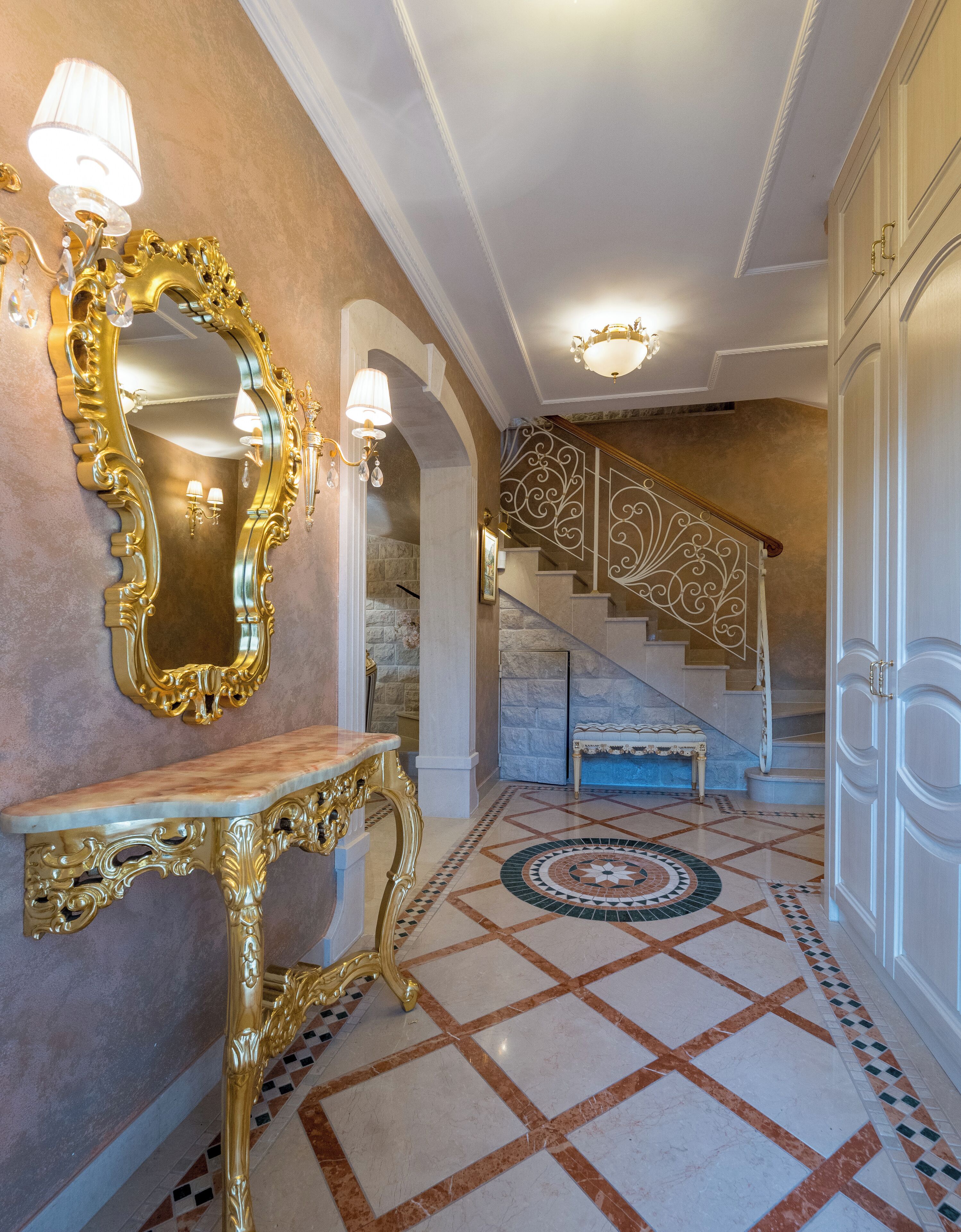 Un amplio pasillo con suelo de mármol, un espejo grande y ornamentado, y una escalera con barandilla decorativa.