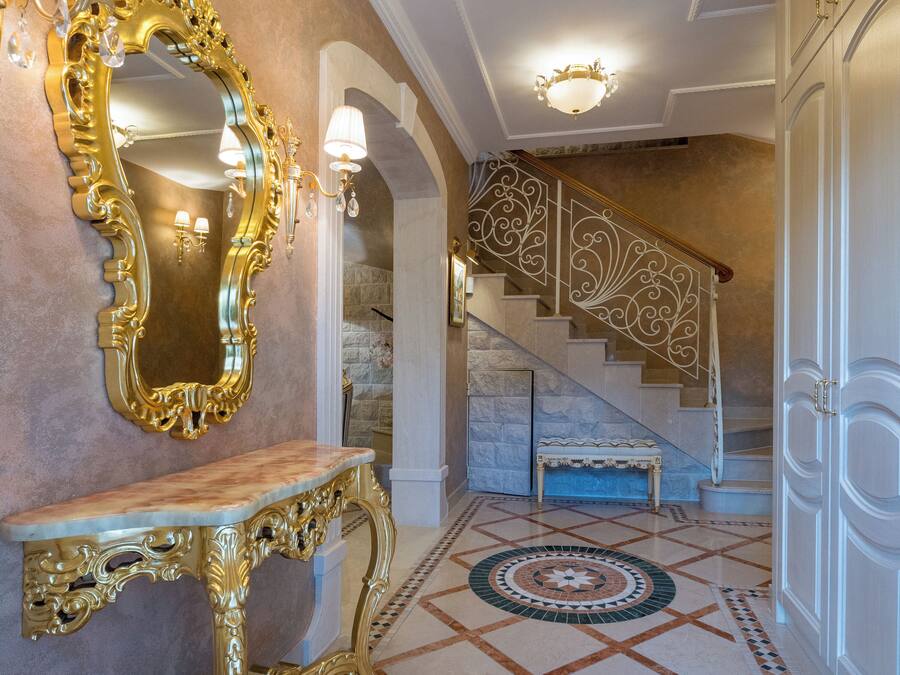 Un amplio pasillo con suelo de mármol, un espejo grande y ornamentado, y una escalera con barandilla decorativa.