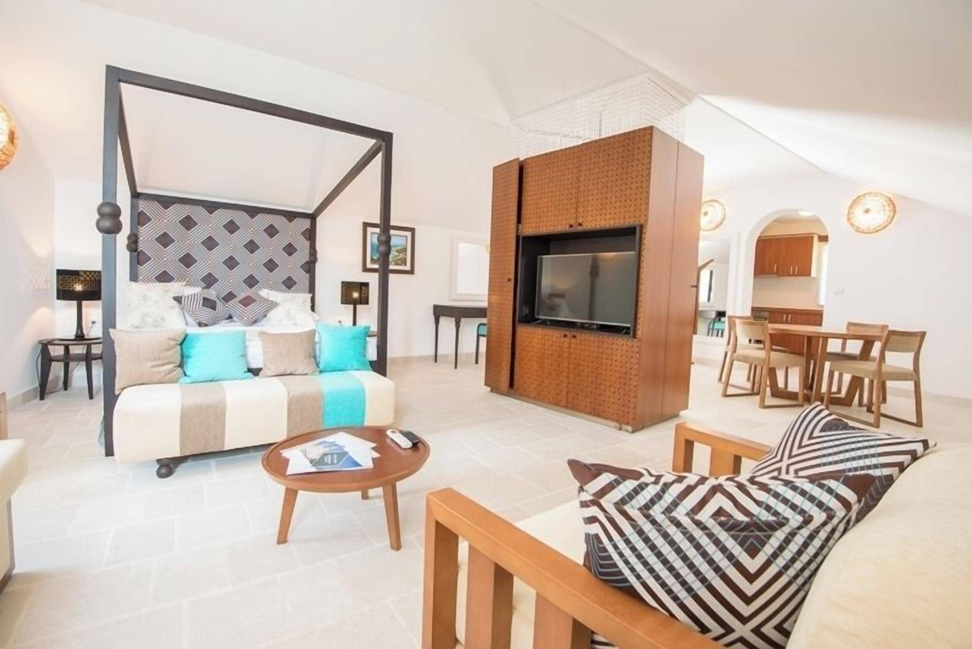 A modern living room with a four-poster bed, a flat-screen TV mounted on a brick wall, and a dining area with wooden furniture.