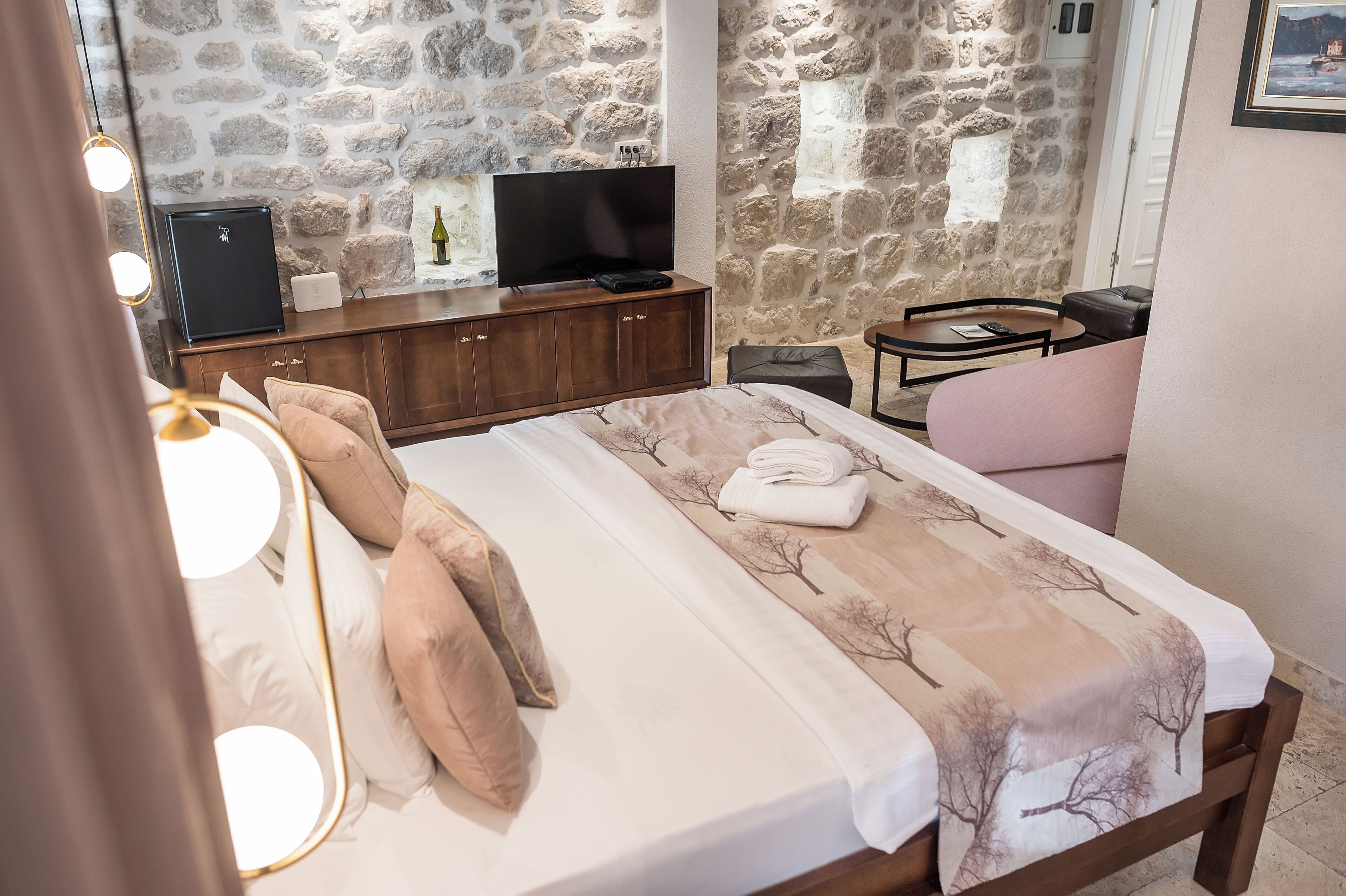A bedroom with a stone wall, a bed with a white and beige bedspread, a wooden dresser with a TV, and a small round table with a chair.
