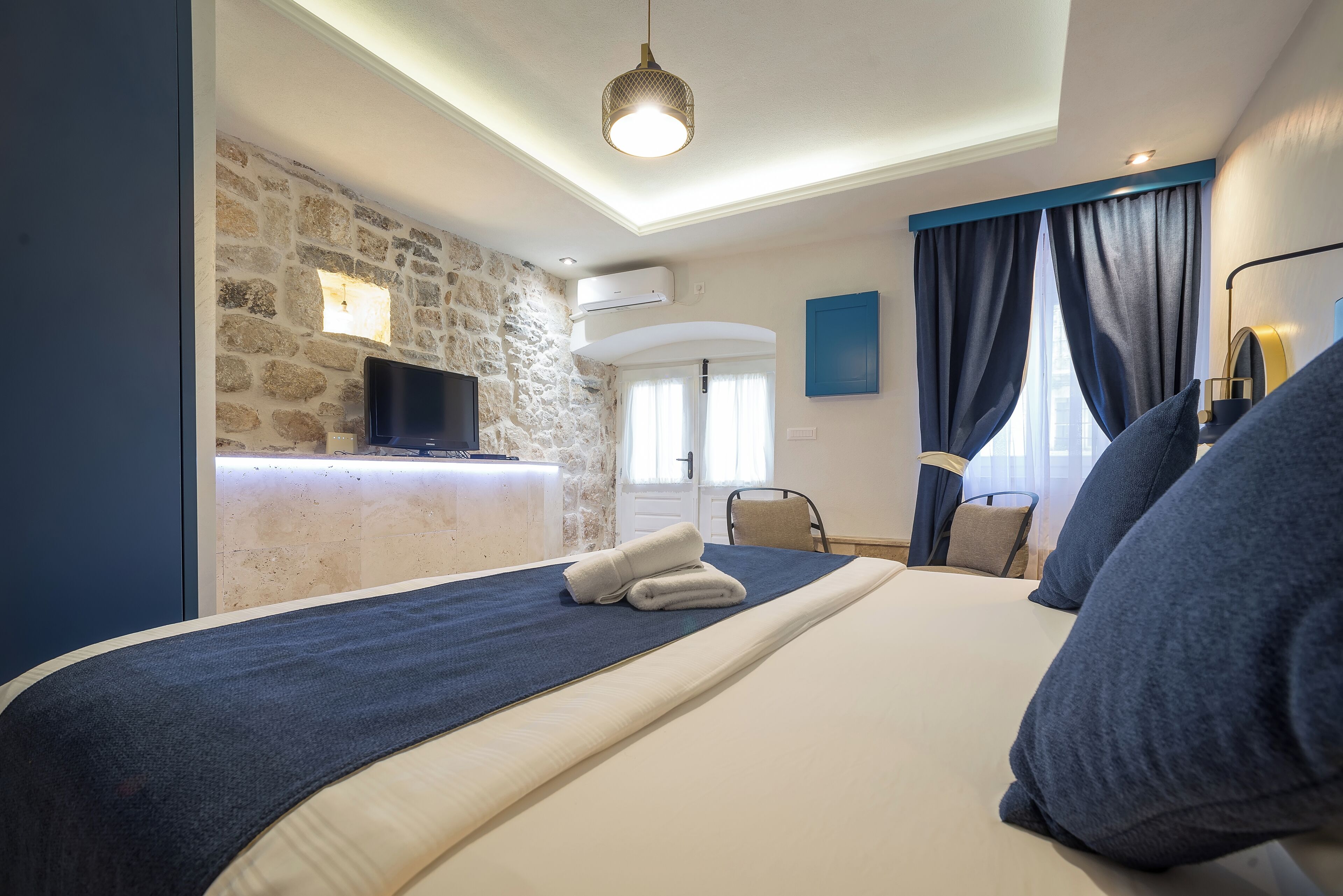 Une chambre d’hôtel avec un mur en pierre, un téléviseur à écran plat, un lit avec une tête de lit bleue et un bureau avec une chaise.