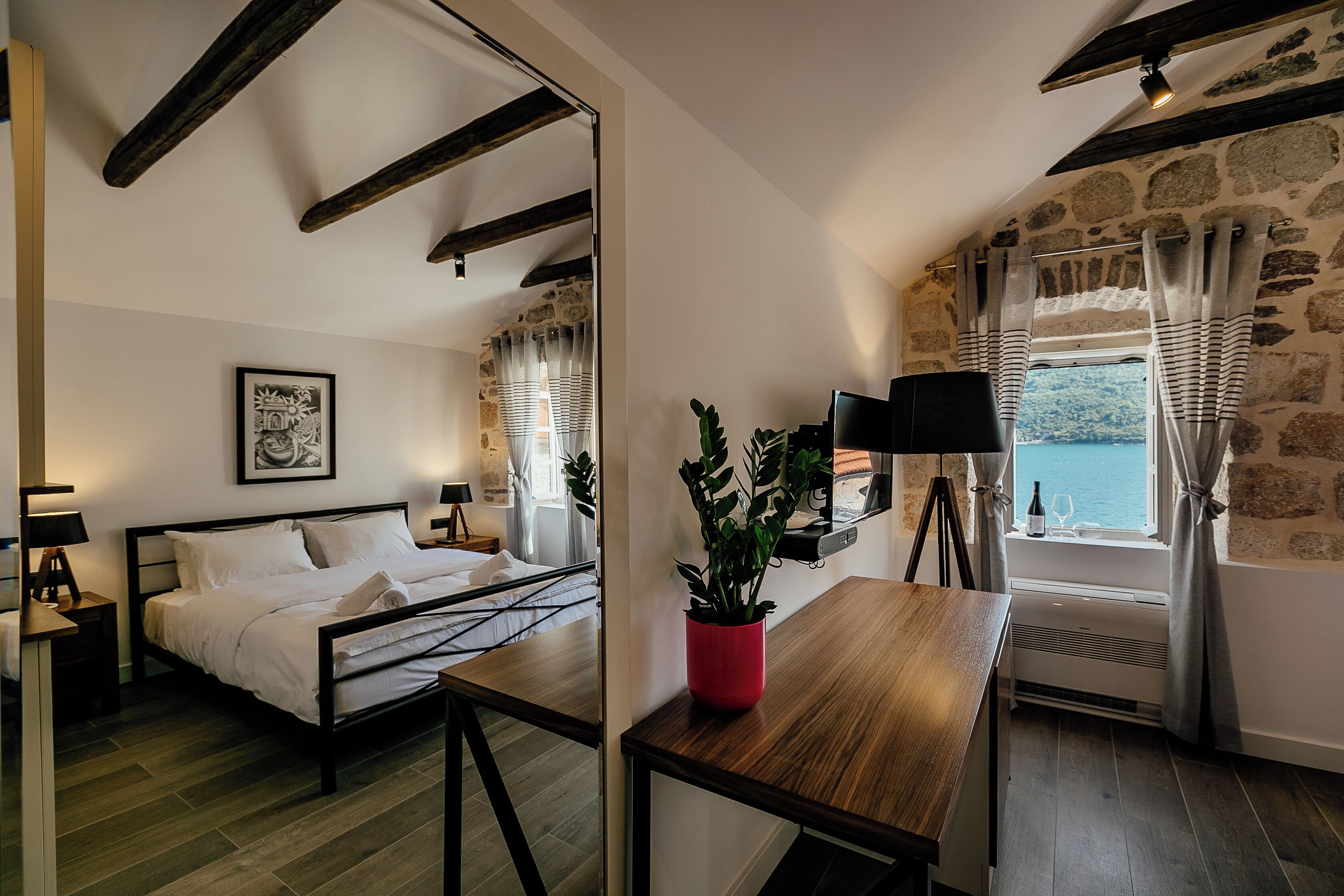 A bedroom with a bed, bedside tables, a red plant, a window with curtains, and a stone wall.