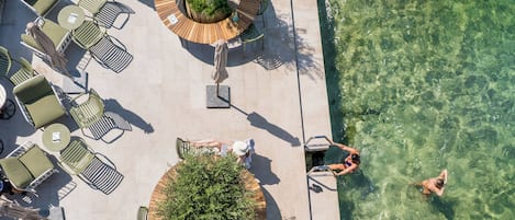 An aerial view of a poolside area with lounge chairs and tables, a clear pool, and a person swimming.