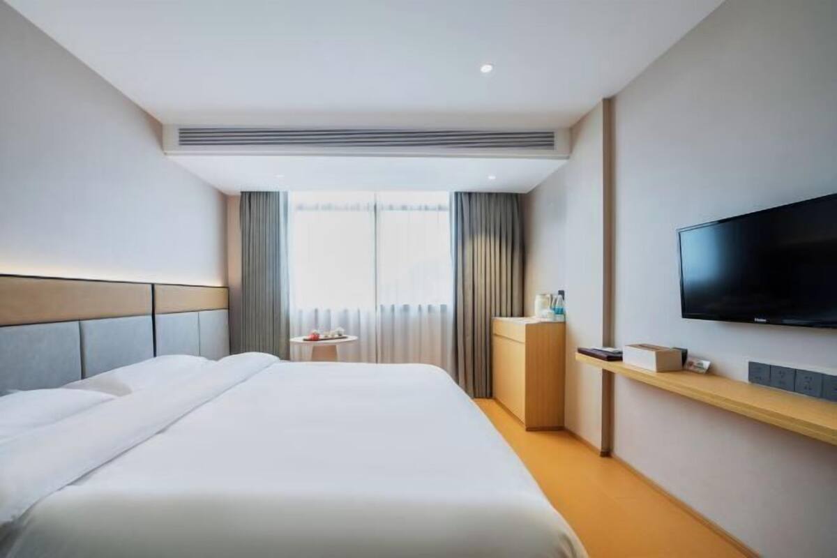 Una habitación de hotel moderna con una cama grande, un televisor de pantalla plana montado en la pared, una mesita de noche pequeña y una ventana con cortinas.
