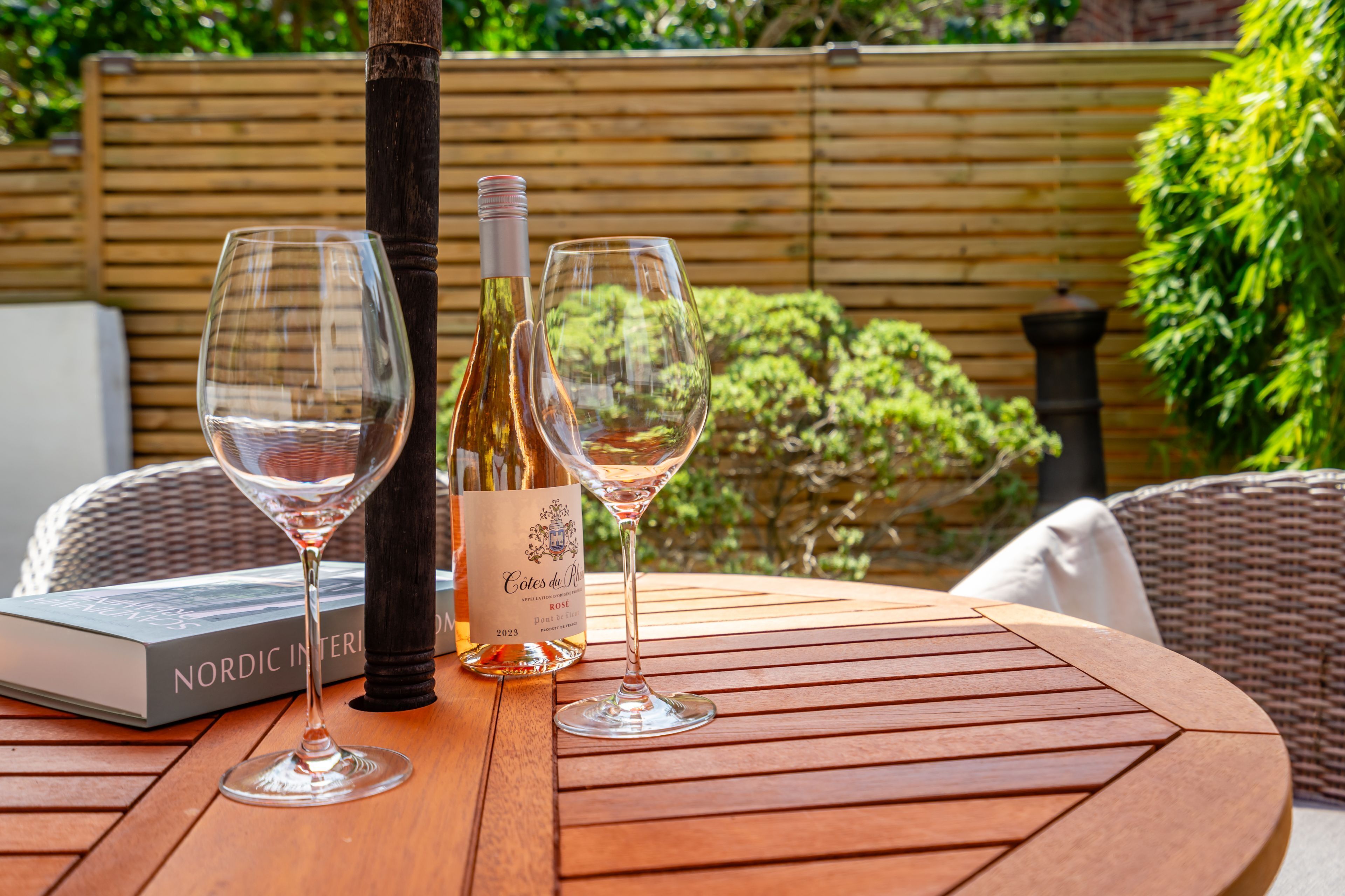 A round wooden table with two wine glasses and a bottle of wine, a book titled 'Nordic Interiors', and a wicker chair.