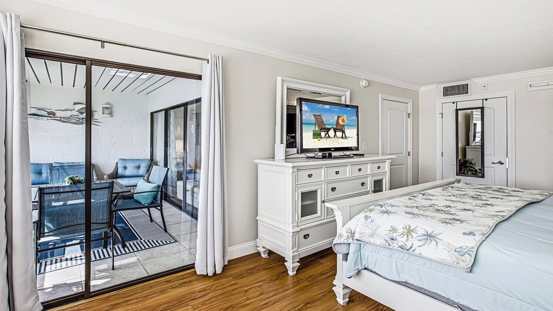 A bedroom with a bed, a television, a dresser, and a sliding glass door leading to a balcony with a table and chairs.