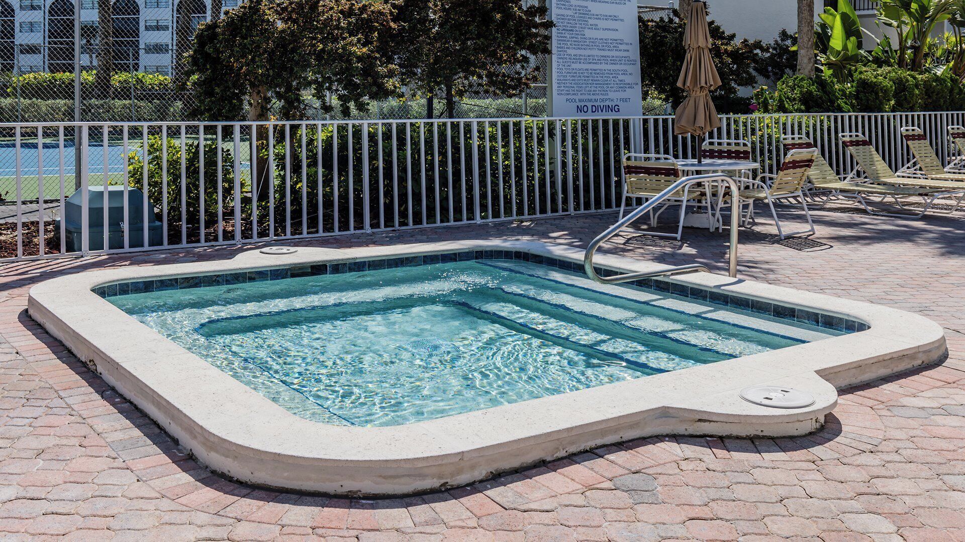 Una piscina rettangolare con scala di sicurezza, circondata da una recinzione in metallo bianco e un cartello che indica una profondità massima di 2,1 metri.