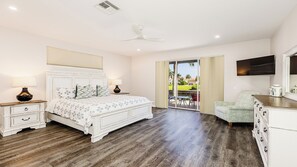 A bedroom with a large bed, a ceiling fan, a TV, and a view of the outdoors.