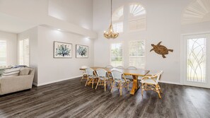 A spacious dining area with a wooden table and wicker chairs, surrounded by white walls and large windows with blinds.