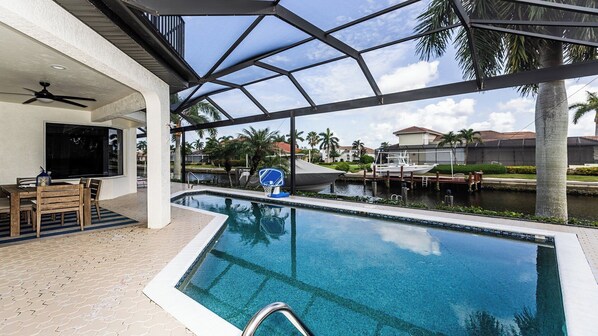 A pool area with a basketball hoop, a seating area with a TV, and a view of a water body and palm trees.