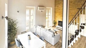 Een ruime woonkamer met een hoge plafond, een trap en een eethoek met een witte tafel en grijze stoelen.