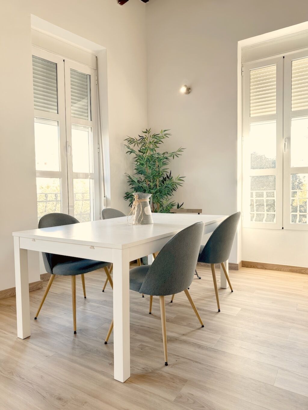 Un comedor con una mesa blanca, cuatro sillas grises, una planta en maceta y un aplique de pared.