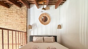 Een slaapkamer met een houten plafond, een bakstenen muur, een bed met beige beddengoed, twee kussens en een ronde wanddecoratie.