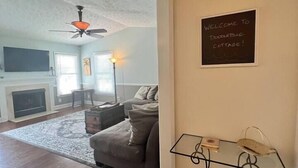 A cozy living room with a sofa, a fireplace, and a ceiling fan. A chalkboard sign reads "Welcome to Doodlebug Cottage!"