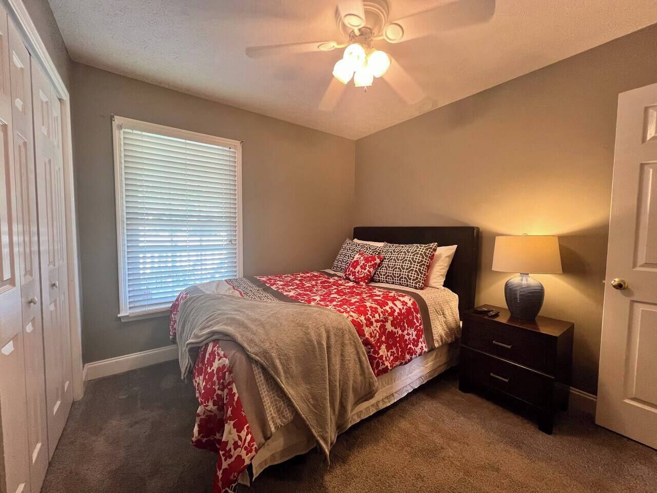 A bedroom with a bed, a nightstand, a lamp, a ceiling fan, a window with blinds, and a door.