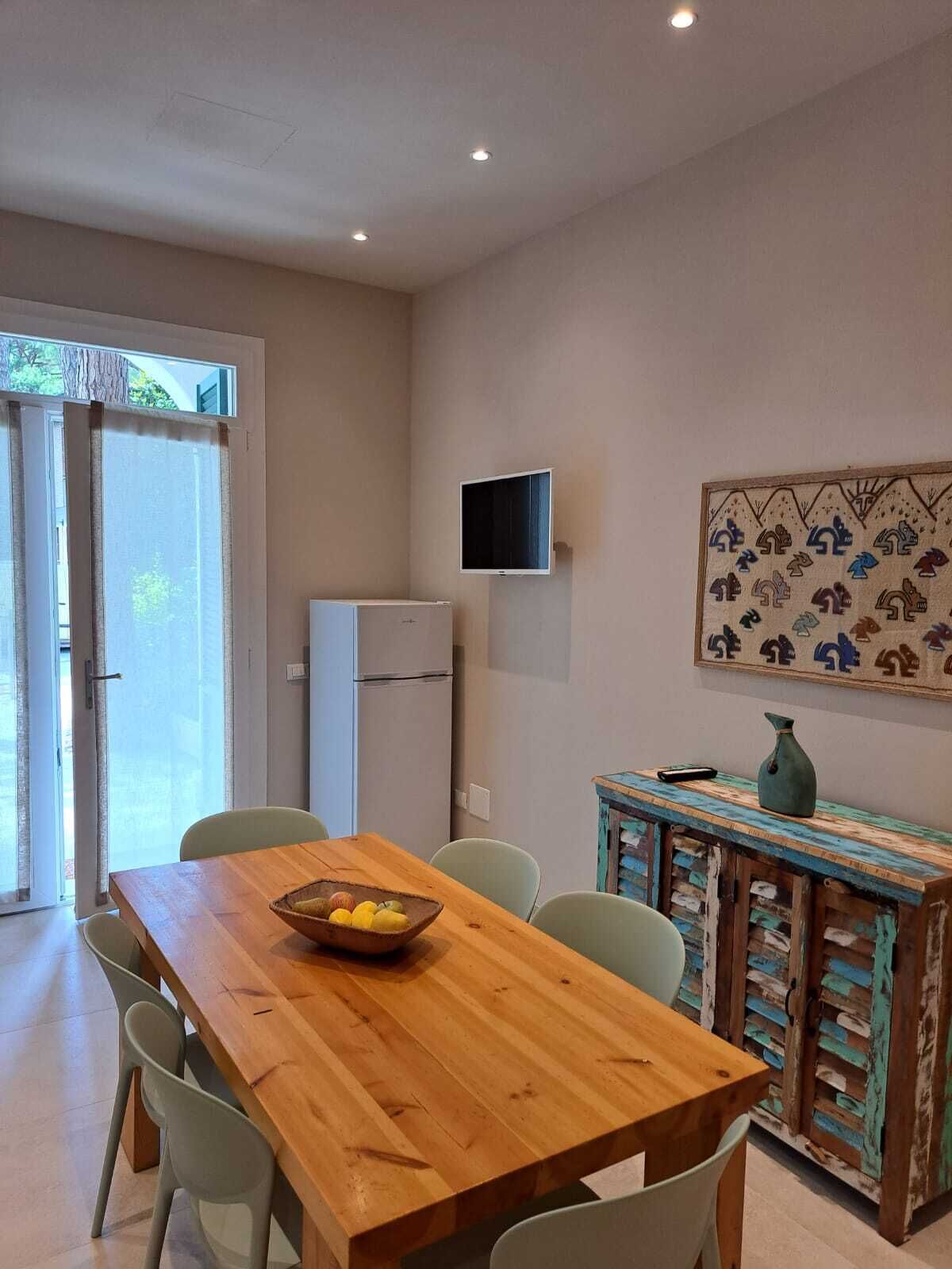 A dining area with a wooden table, chairs, a refrigerator, a television mounted on the wall, and a colorful cabinet.