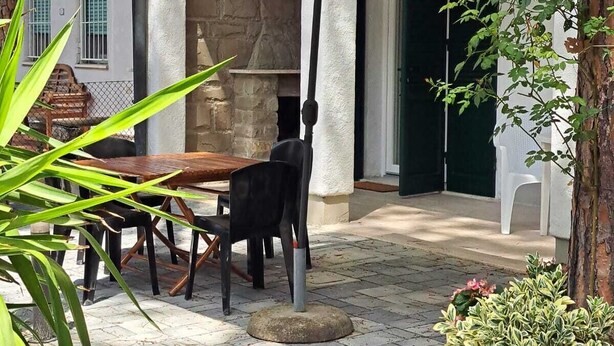 An outdoor patio with a table and chairs under a white umbrella, surrounded by greenery and a stone wall. - Marina Romea 2 Splitted Executive Flats (Ravenna)