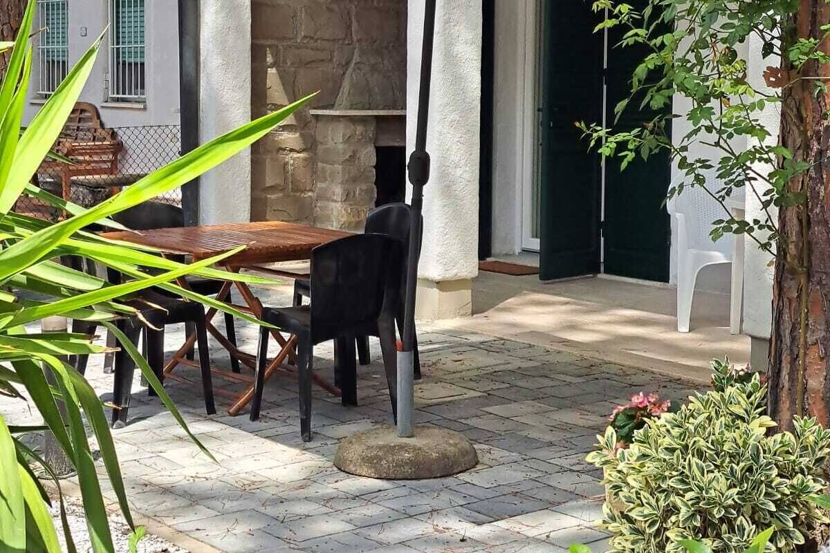 An outdoor patio with a table and chairs under a white umbrella, surrounded by greenery and a stone wall.