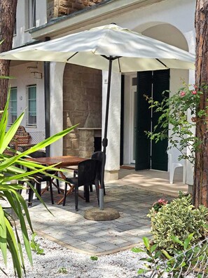 Um pátio externo com mesa e cadeiras sob um guarda-sol branco, cercado por vegetação e uma parede de pedra.