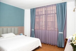 Quarto de hotel com cama, mesa de cabeceira, janela com cortinas leves e um vaso com flores.