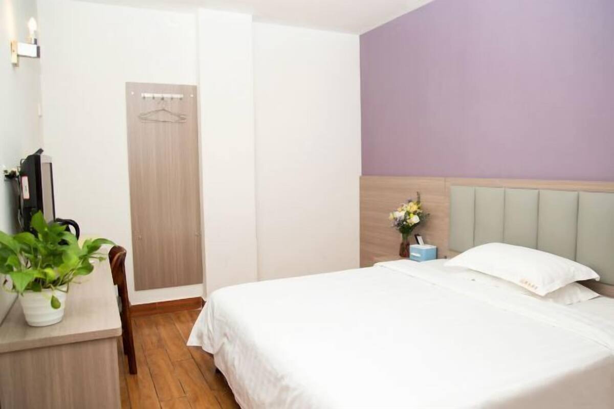 A bedroom with a bed, a bedside table with a plant, a nightstand, a wardrobe, and a hanging garment rack.