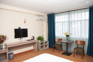 Quarto de hotel com televisão de tela plana, uma escrivaninha, duas cadeiras, uma mesinha com um vaso de flores e uma janela grande com cortinas.
