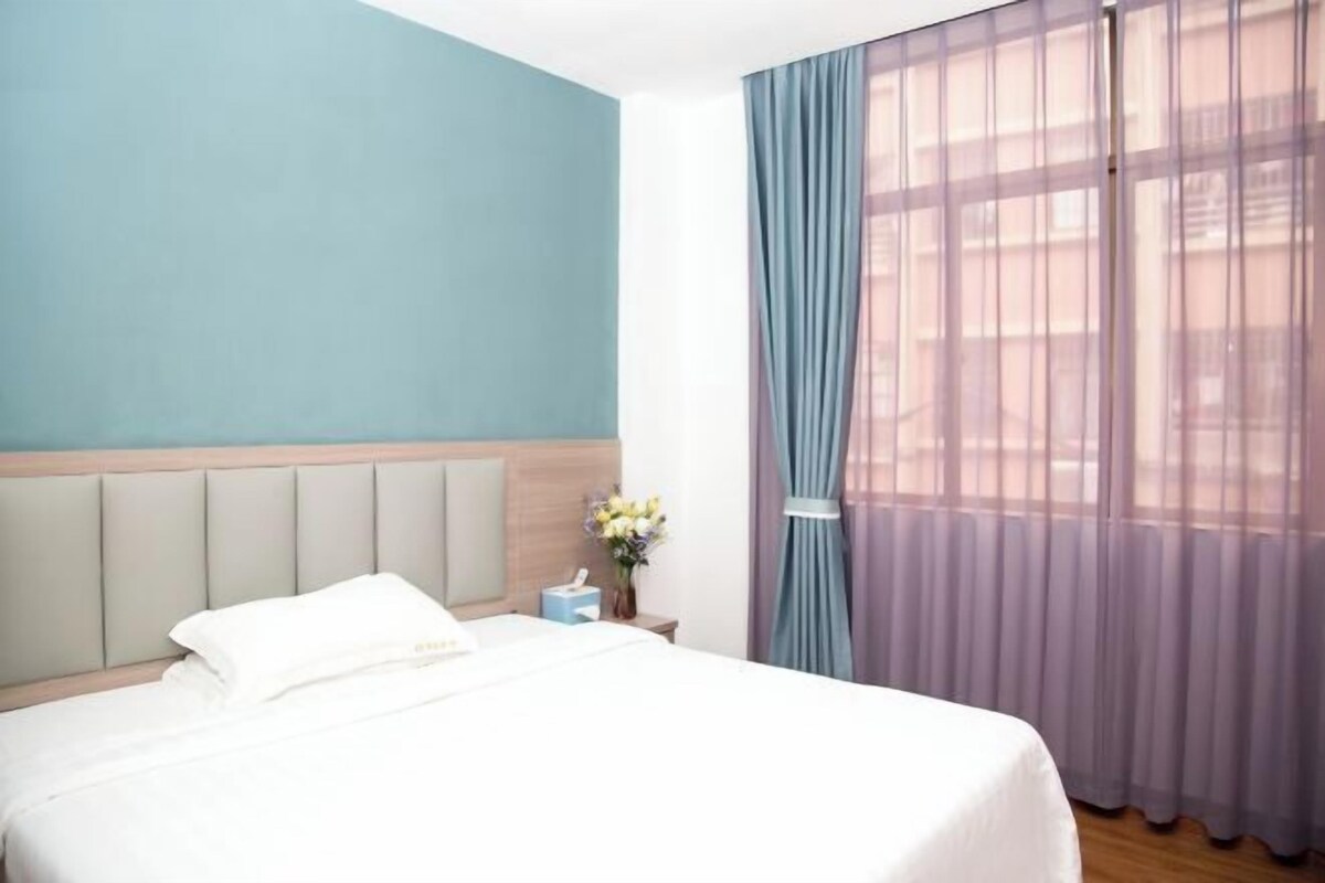 Quarto de hotel com cama, mesa de cabeceira, vaso de flores e janela com cortinas leves.