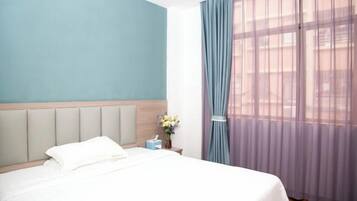 Quarto de hotel com cama, mesa de cabeceira, vaso de flores e janela com cortinas leves.
