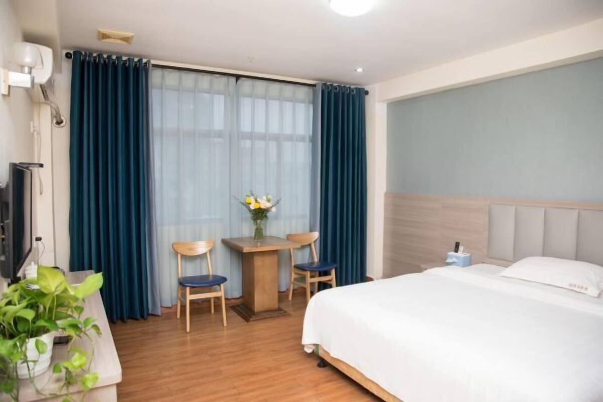 A hotel room with a bed, a desk with two chairs, a small table with a vase of flowers, and a television on the left side.