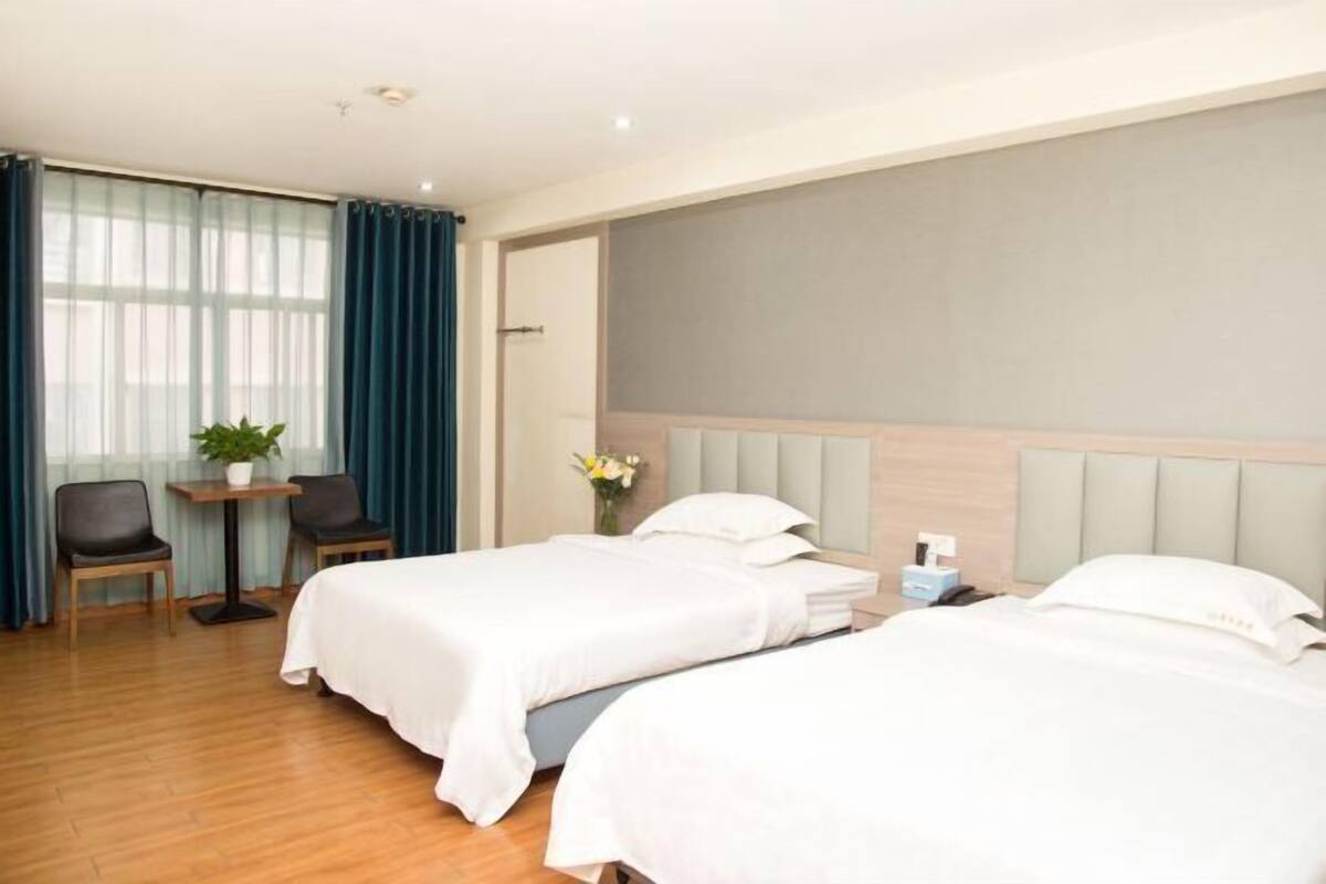 A hotel room with two beds, a small table with chairs, a potted plant, and a window with curtains.