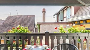 Een balkon met een tafel voor twee personen, voorzien van een rode tafelkleed met een hartmotief, twee stoelen met blauwe kussens en een uitzicht op een gebouw met een pannendak en schoorsteen.