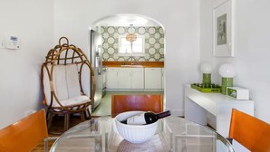 A dining area with a glass table, wicker chairs, a wine bottle, and a kitchen in the background with patterned wallpaper.