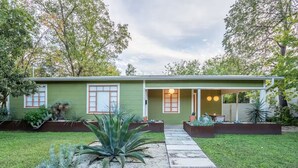 A modern house with a green exterior, two windows with white frames, a covered porch with a table and chairs, and a well-maintained lawn with various plants.