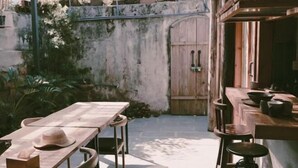 Un espacio exterior rústico para comer, con mesa y sillas de madera, un mostrador de madera con taburetes y una pared de concreto con una puerta de madera.