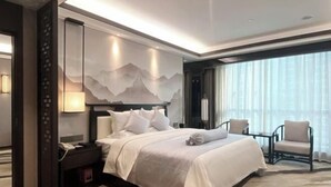 Ein modernes Hotelzimmer mit einem großen Bett, einem Nachttisch, einem Sessel und Blick auf die Stadt durch große Fenster.