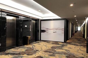Pasillo de hotel con alfombra estampada, puertas de ascensor negras y una pantalla grande montada en la pared mostrando un paisaje.