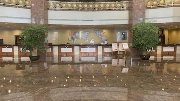 A hotel lobby with a marble floor, reception desk, two large potted plants, and a world map on the wall.