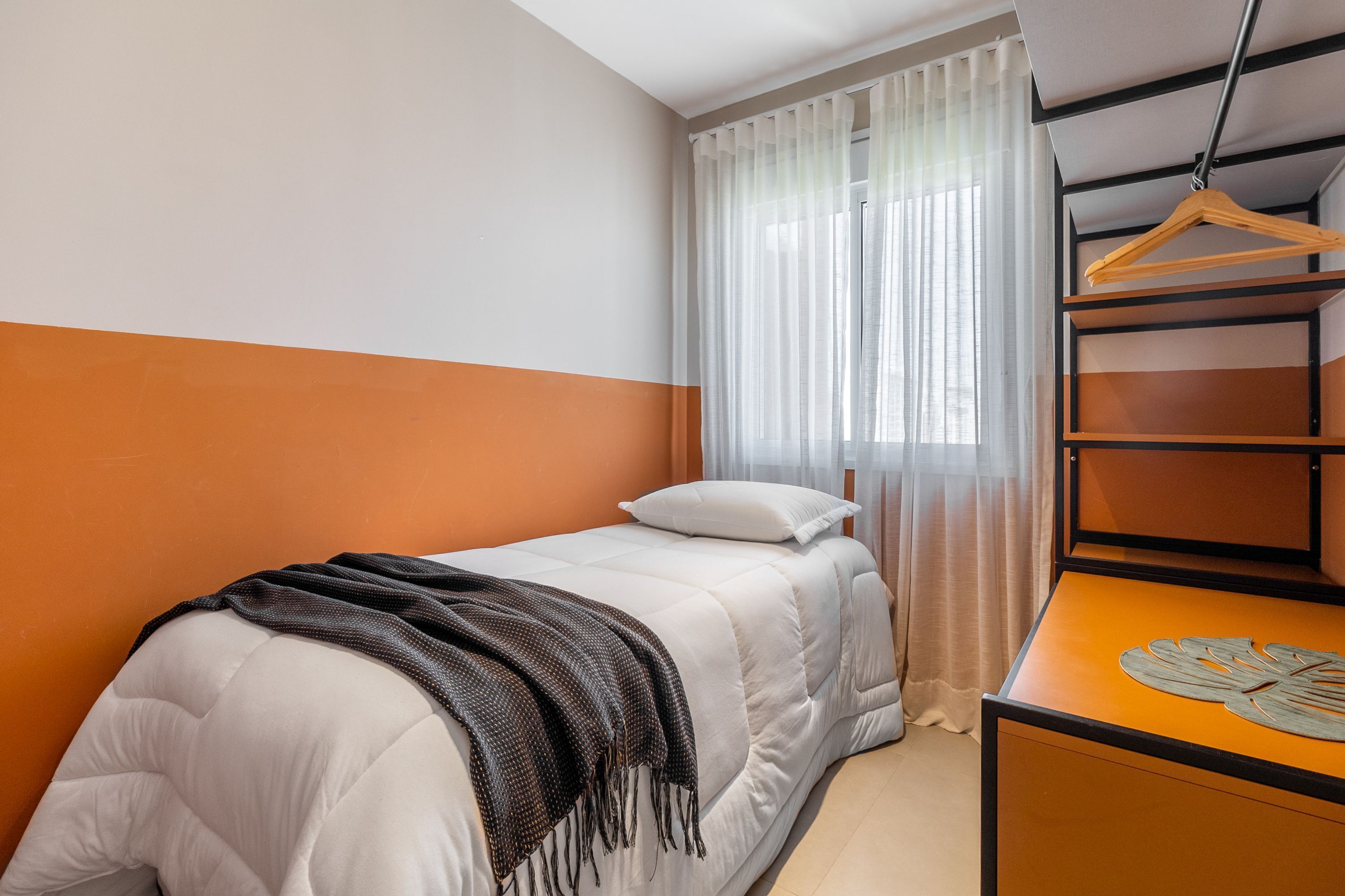 A single bed with a white headboard and a brown blanket, a wooden dresser with a leaf-shaped decoration, and a hanging wooden hanger.