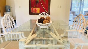 A dining area with a glass table surrounded by white chairs with woven bases, a decorative basket with woven balls, and a red painting on the wall.