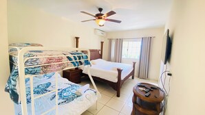 A hotel room with a bunk bed, a single bed, a wooden nightstand, a small table with electronic devices, a ceiling fan, and a window with curtains.
