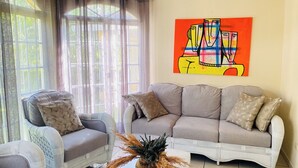 A living room with a grey sofa, a wicker armchair, and a large window with sheer curtains.