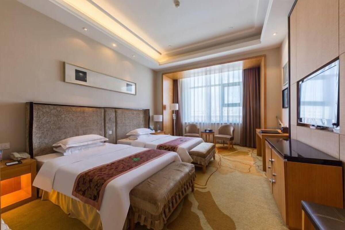 Habitación de hotel con dos camas, un escritorio con televisor, una silla y un ventanal con cortinas.