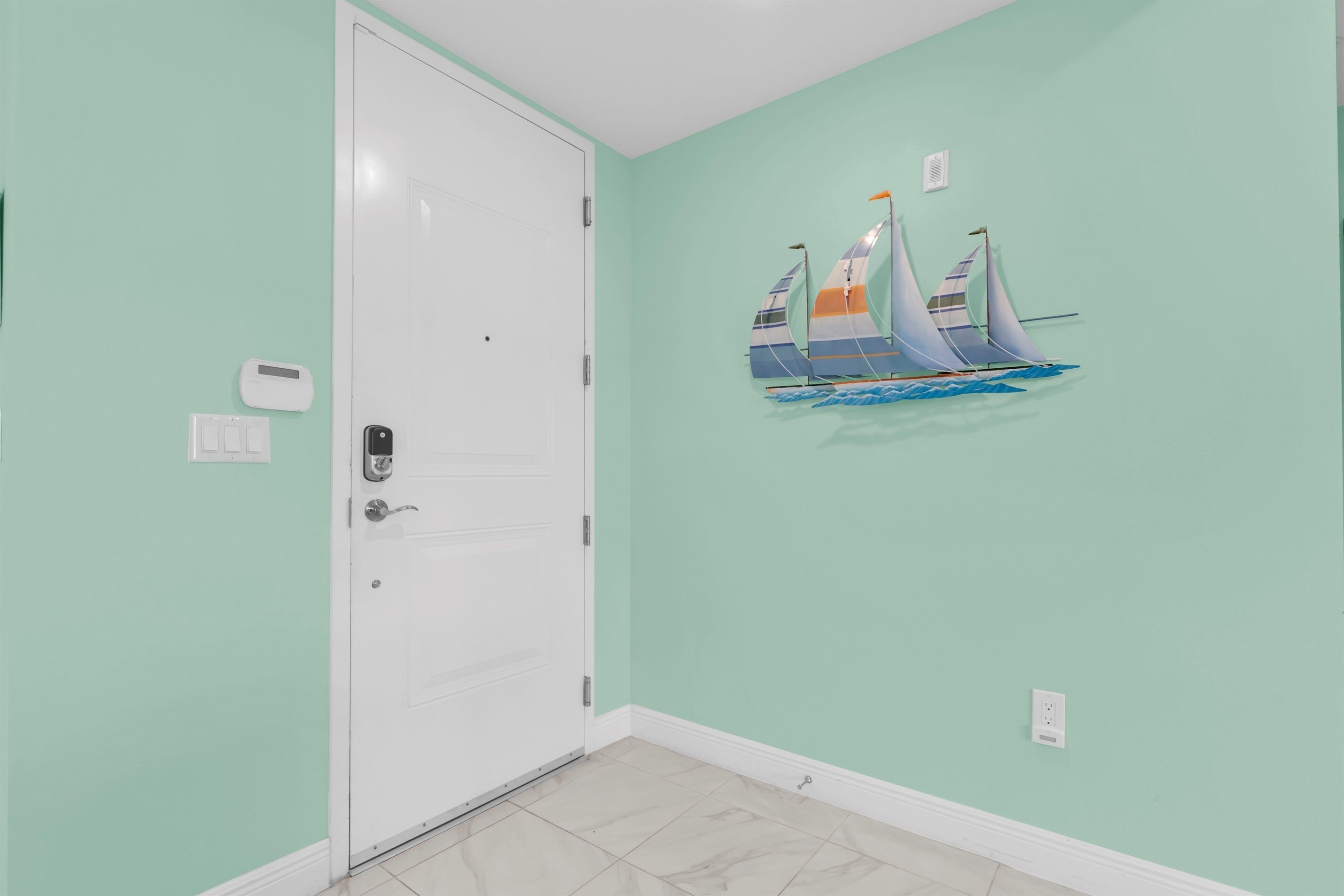 A room with a white door, a wall-mounted thermostat, a wall-mounted light switch, and a wall decoration of sailboats.