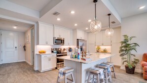 Una cocina moderna con armarios blancos, una isla central con taburetes, electrodomésticos de acero inoxidable e iluminación colgante.