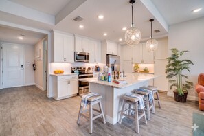 A modern kitchen with white cabinetry, a central island with stools, stainless steel appliances, and pendant lighting. - Sunset Seacret by Coastal Chic Vacations (Bradenton)