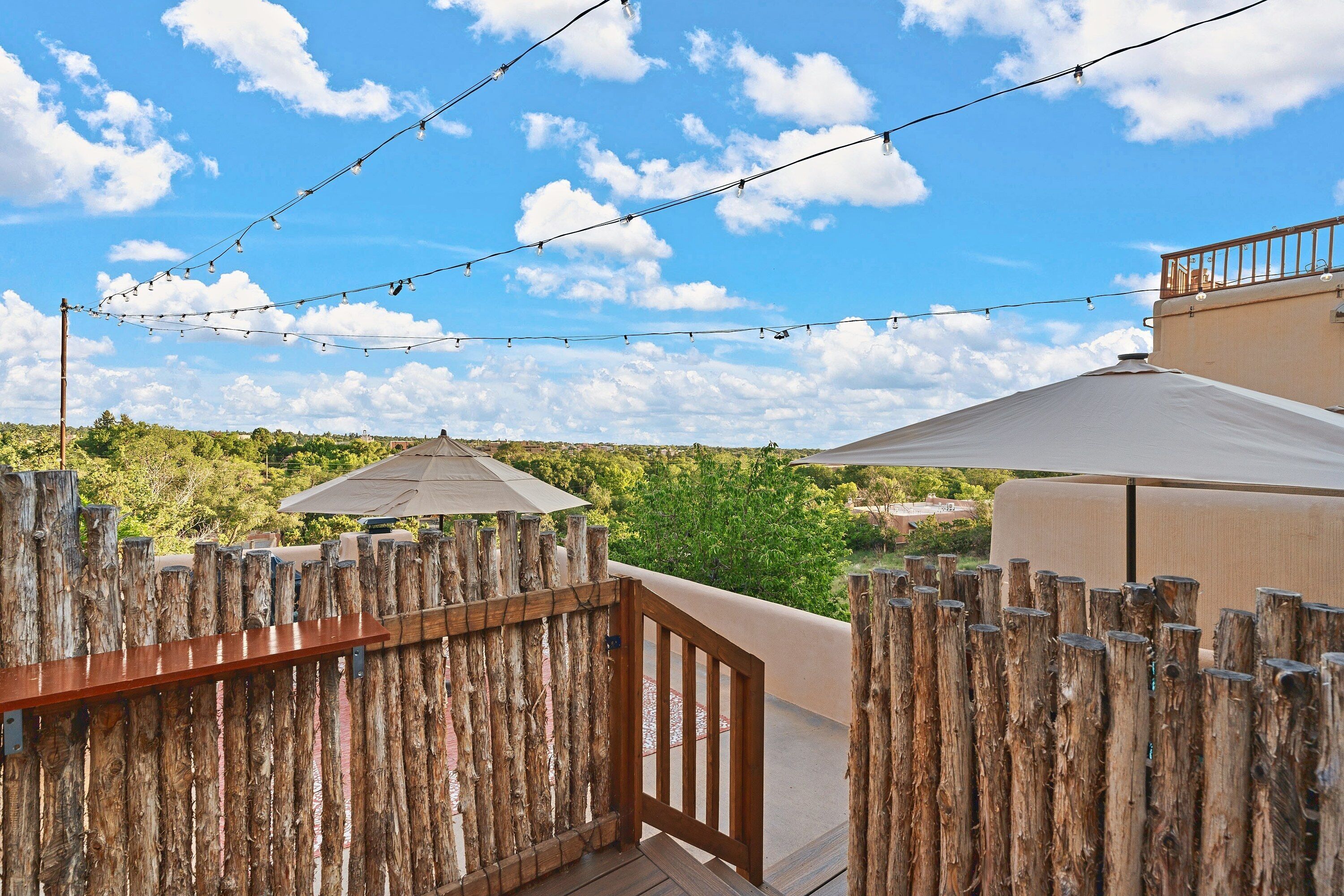 A wooden deck with a rustic fence, a pergola with a canopy, and string lights against a backdrop of a clear sky and greenery.